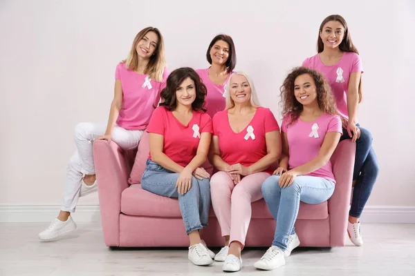 Group of women with silk ribbons on sofa near light wall. Breast cancer awareness concept