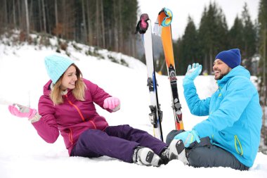 Happy couple with ski equipment sitting on snowy hill in mountains, space for text. Winter vacation