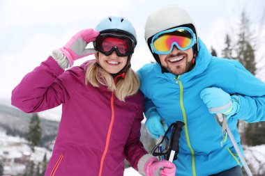 Happy couple with ski equipment sitting on snowy hill in mountains, space for text. Winter vacation