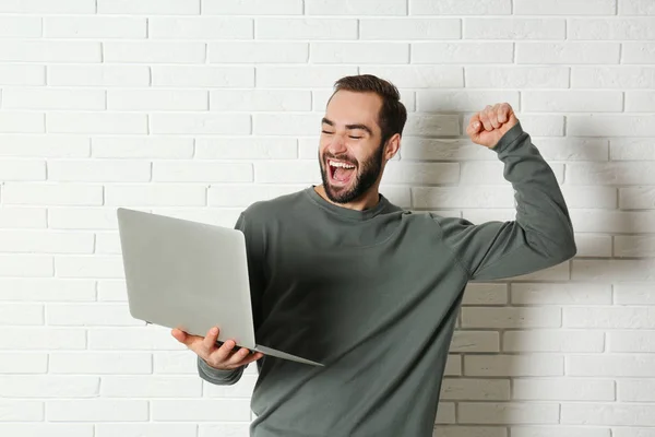 Emotional young man with laptop celebrating victory near brick wall