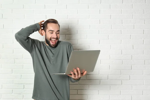 Emotional young man with laptop celebrating victory near brick wall. Space for text