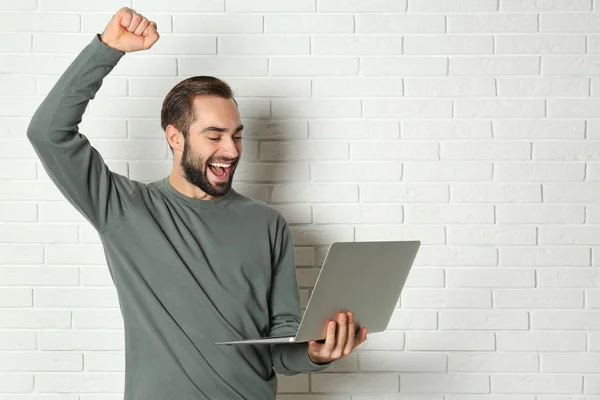 Emotional young man with laptop celebrating victory near brick wall. Space for text