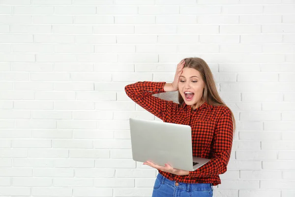 Emotional young woman with laptop celebrating victory near brick wall. Space for text