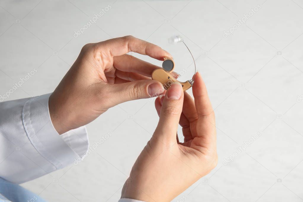 Woman putting battery into hearing aid on light background, closeup