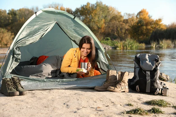 Young woman having breakfast in sleeping bag inside of camping tent