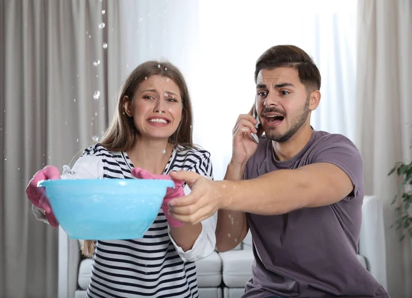 Emotional young woman collecting water leaking from ceiling while her husband calling plumber in living room