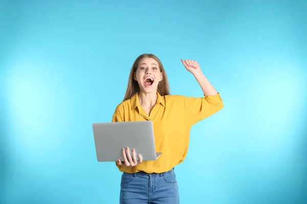 Emotional young woman with laptop celebrating victory on color background