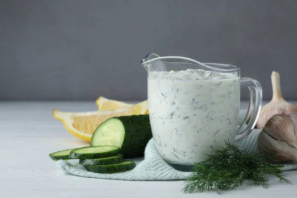 Glass jug of Tzatziki cucumber sauce with ingredients on grey background, space for text
