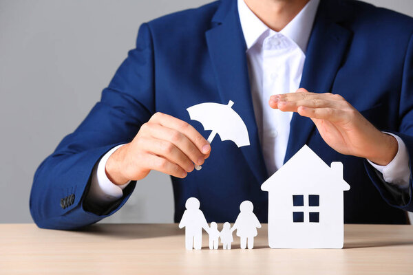 Male insurance agent covering paper family and home with umbrella cutout at table, closeup