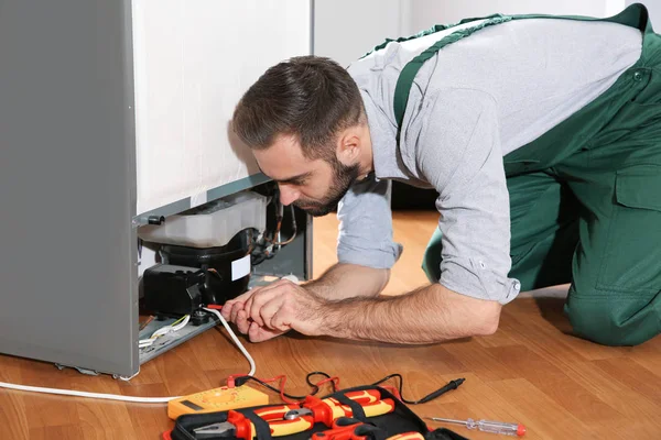 Male technician in uniform repairing refrigerator indoors
