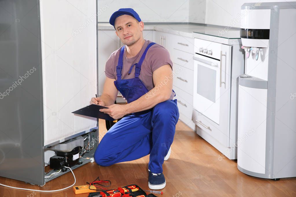 Male technician with clipboard and tools near broken refrigerator in kitchen