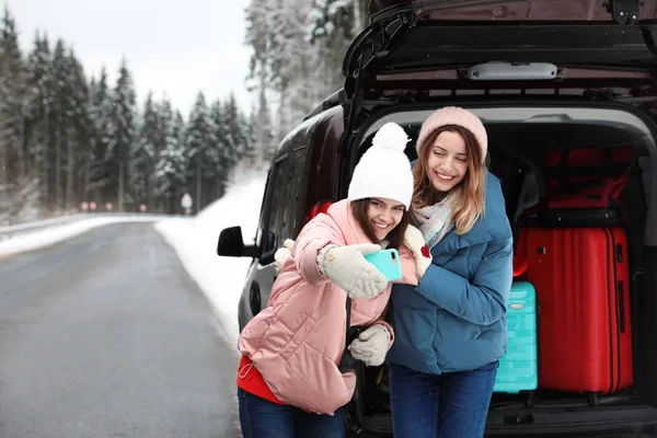 Friends taking selfie near open car trunk full of luggage on road, space for text. Winter vacation
