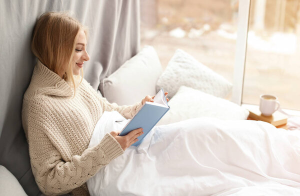 Красивая молодая женщина в вязаном свитере сидит и читает книгу у окна дома. Зимняя атмосфера
