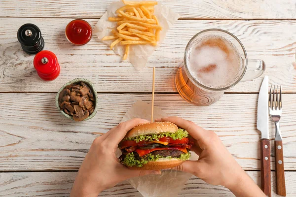 Woman holding tasty burger with bacon over wooden table, top view