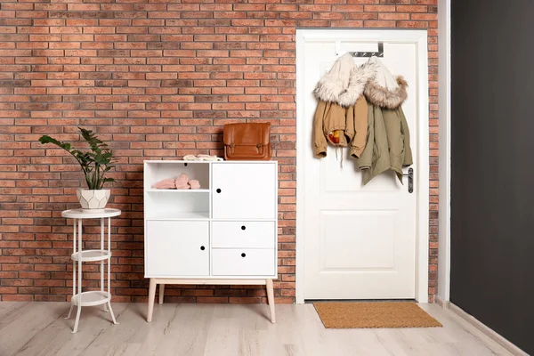 Stylish hallway interior with chest of drawers and clothes hanging on white door