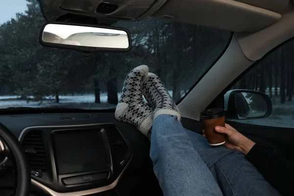 Young man in warm socks with coffee resting inside car, closeup