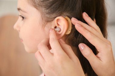 Young woman putting hearing aid in daughter's ear indoors, closeup clipart