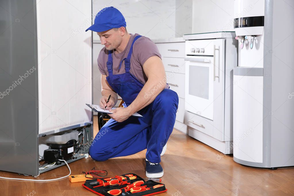 Male technician with clipboard examining broken refrigerator in kitchen