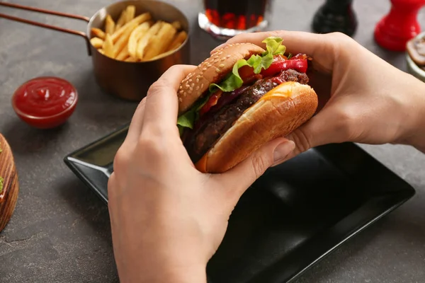 Woman holding tasty burger with bacon over grey table, closeup