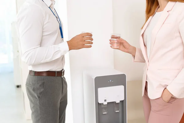 Co-workers having break near water cooler at workplace, closeup