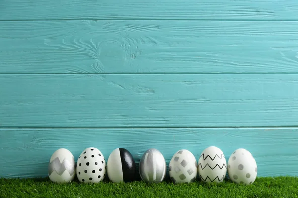 Line of painted Easter eggs on green lawn against wooden background, space for text