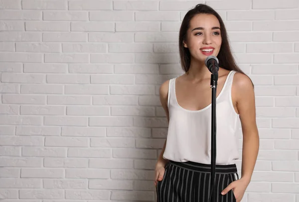 Young woman in casual clothes posing with microphone near brick wall. Space for text