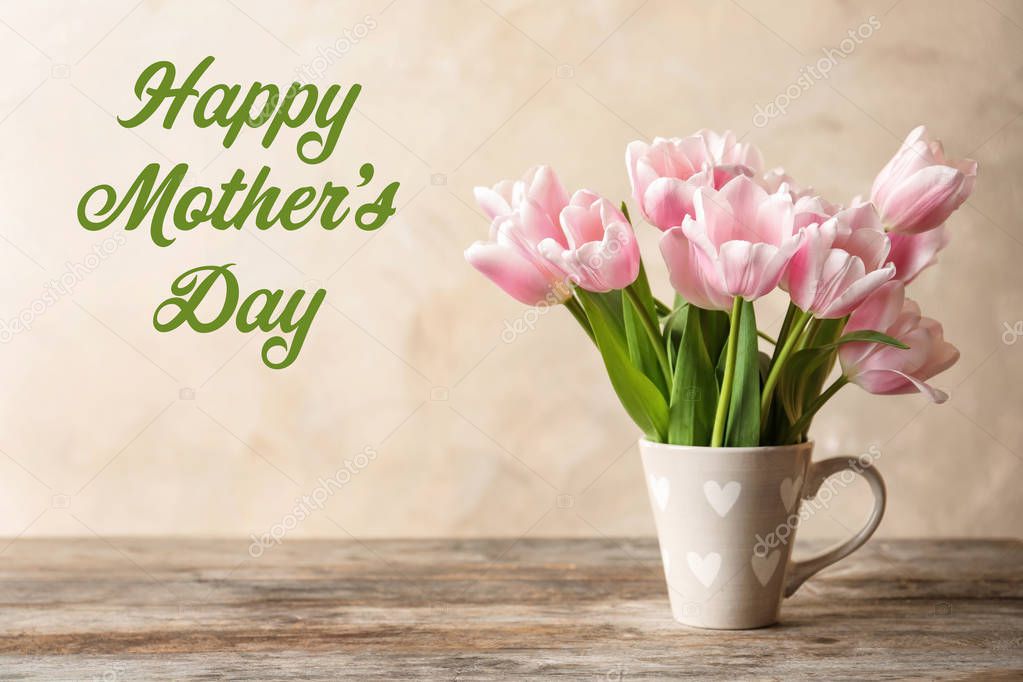 Beautiful tulips in cup on table and text Happy Mother's Day against color background