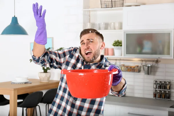 Emotional young man calling plumber while collecting water leakage from ceiling in kitchen