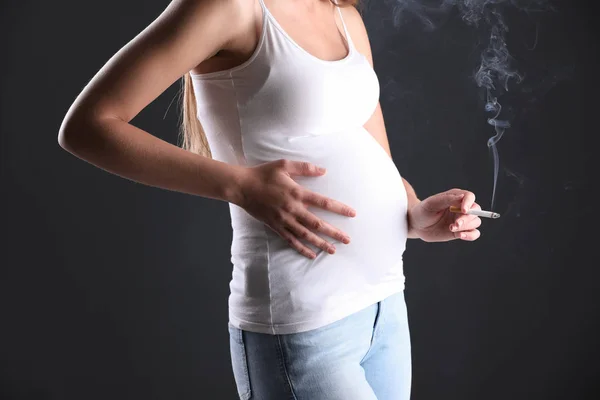 Young pregnant woman smoking cigarette on dark background, closeup. Harm to unborn baby