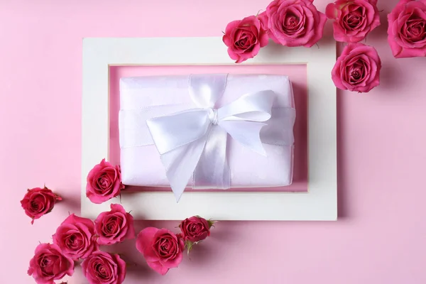 Frame with roses and gift box on color background, top view