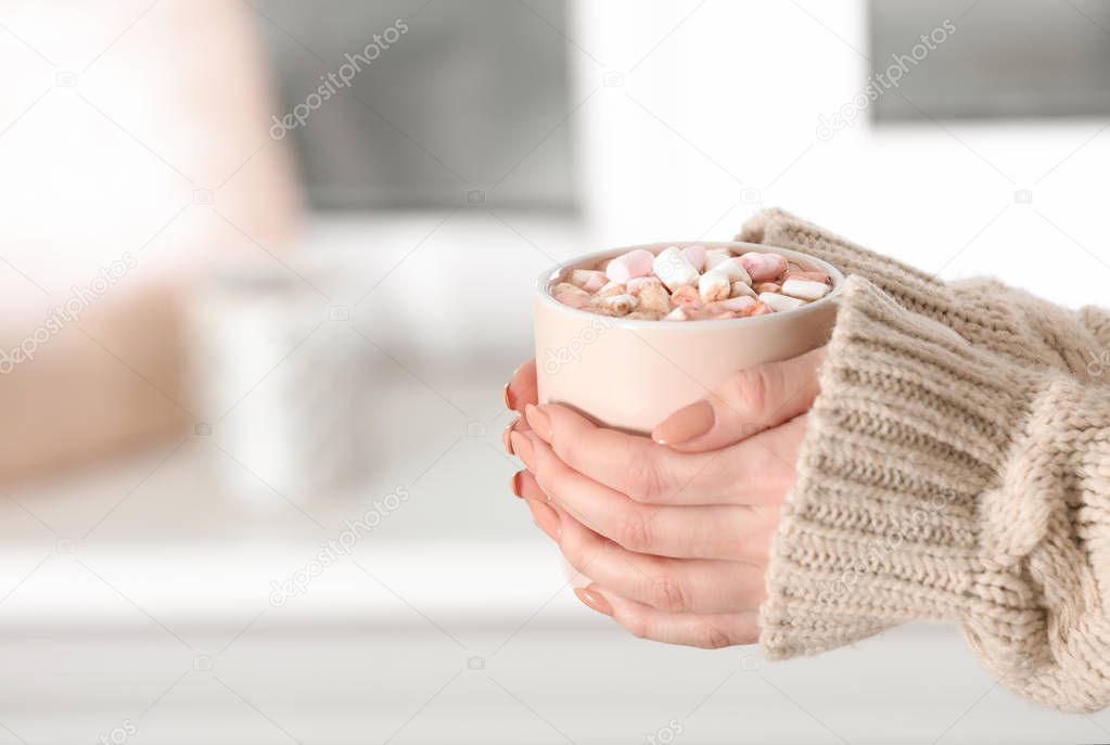 Woman holding cup of aromatic cacao with marshmallows on blurred background, closeup. Space for text