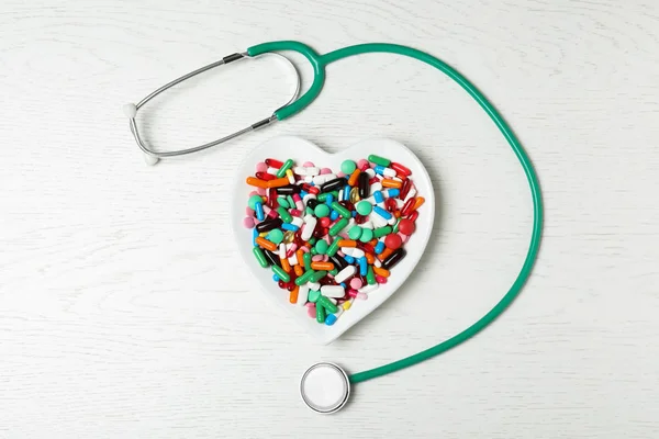 Plate with heart pills and stethoscope on wooden background, flat lay. Cardiology concept