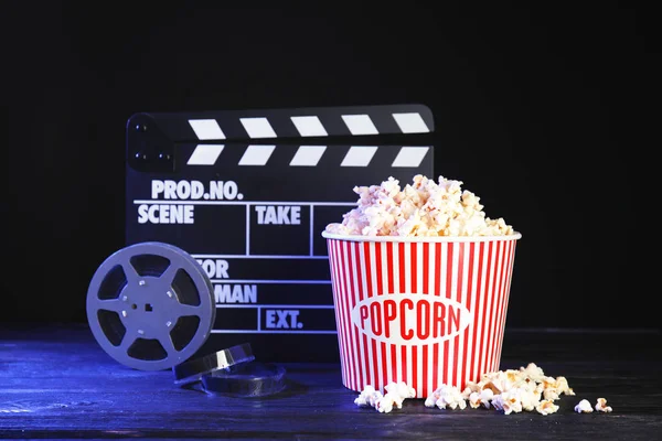 Clapperboard, reel and popcorn bucket on table against dark background. Watching cinema