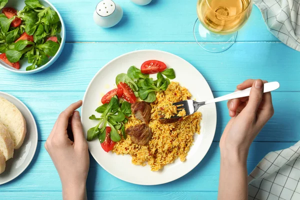 Woman eating rice pilaf with meat and salad at table, top view