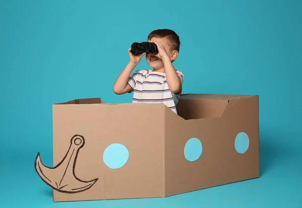 Lindo niño jugando con prismáticos y barco de cartón sobre fondo de color — Foto de Stock