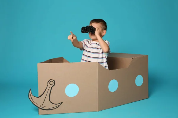 Lindo niño jugando con prismáticos y barco de cartón sobre fondo de color — Foto de Stock