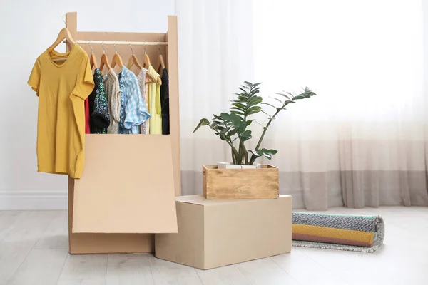 Caixa de guarda-roupa de papelão com roupas em cabides, planta de sala e carpete dentro de casa. Dia de mudança — Fotografia de Stock