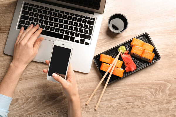 Woman with smartphone, laptop and tasty sushi rolls at wooden table, space for text. Food delivery