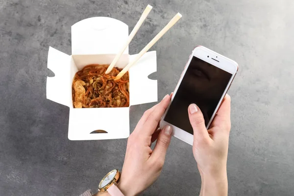 Mulher com smartphone e macarrão chinês na mesa cinza, espaço para texto. Entrega de alimentos — Fotografia de Stock