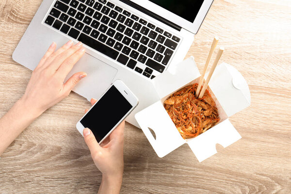 Woman with smartphone, laptop and Chinese noodles at wooden table, space for text. Food delivery