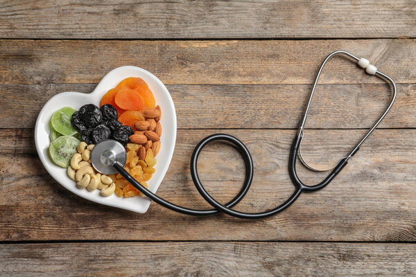 Пластина в форме сердца с орехами и сухофруктами возле стетоскопа на деревянном фоне, вид сверху

