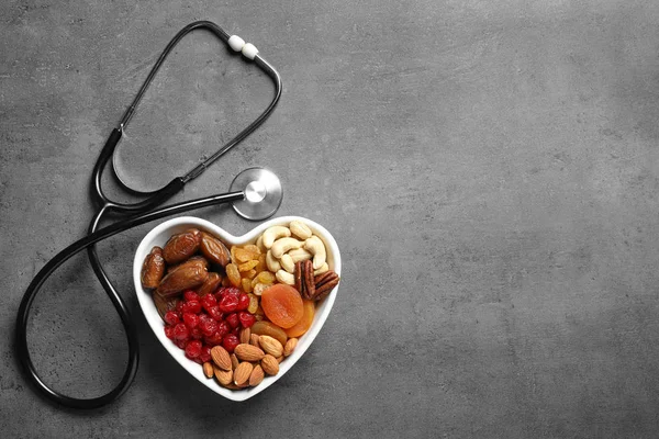 Herzförmige Schale mit Nüssen und Trockenfrüchten in der Nähe von Stethoskop auf grauem Hintergrund, Draufsicht. Raum für Text — Stockfoto
