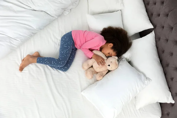 Cute little African-American girl with teddy bear sleeping in bed, top view — Stock Photo, Image