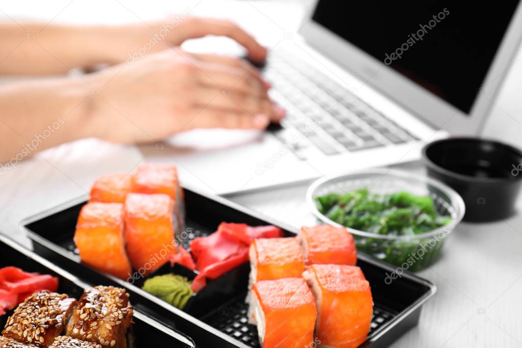 Boxes with different sushi rolls and blurred woman using laptop on background. Food delivery