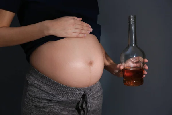 Беременная женщина с бутылкой виски на темном фоне. Алкогольная зависимость — стоковое фото