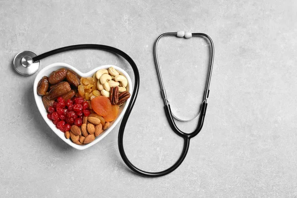 Herzförmige Schale mit Nüssen und Trockenfrüchten in der Nähe von Stethoskop auf grauem Hintergrund, Draufsicht. Raum für Text — Stockfoto