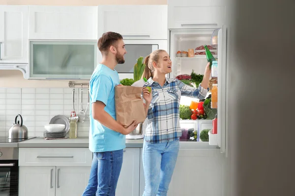 Ευτυχισμένο ζευγάρι με χάρτινη σακούλα γεμάτη προϊόντα στέκεται κοντά ψυγείο στην κουζίνα — Φωτογραφία Αρχείου