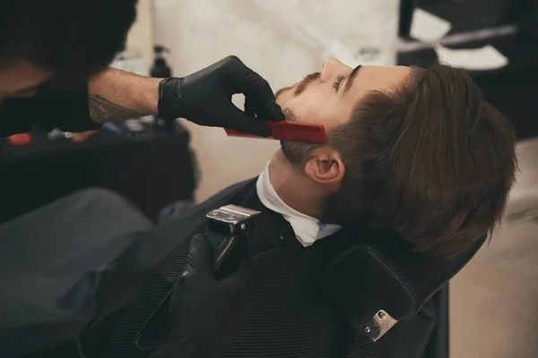 Парикмахер стрижет бороду клиенту в парикмахерской. Профессиональные услуги бритья — стоковое фото