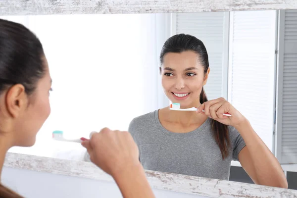 Jovem mulher limpando os dentes contra espelho no banheiro — Fotografia de Stock