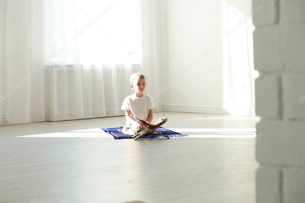 Little Muslim boy with Koran praying on rug indoors